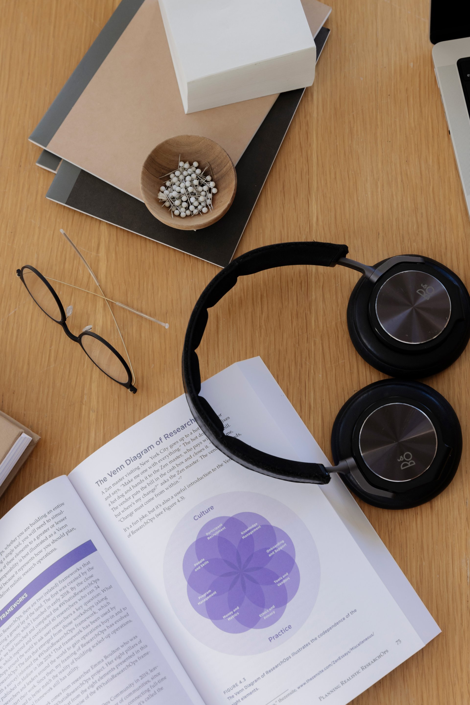 A top down view of a table with a book, Research That Scales The Research Operations Handbook, open and being read. A pair of spectacles and head phones sit atop the table.